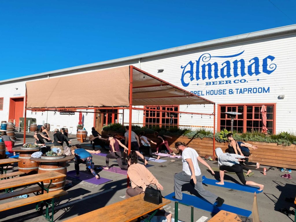 The image depicts the exterior of the Almanac Beer Co. barrel house taproom. The taproom is a large building with a gray exterior and a black roof. The entrance is framed by two large wooden doors and there are several windows along the sides of the building. In front of the taproom, there is a spacious outdoor area with several wooden tables and chairs where customers can sit and enjoy their drinks. The Almanac Beer Co. logo is prominently displayed above the entrance to the taproom.