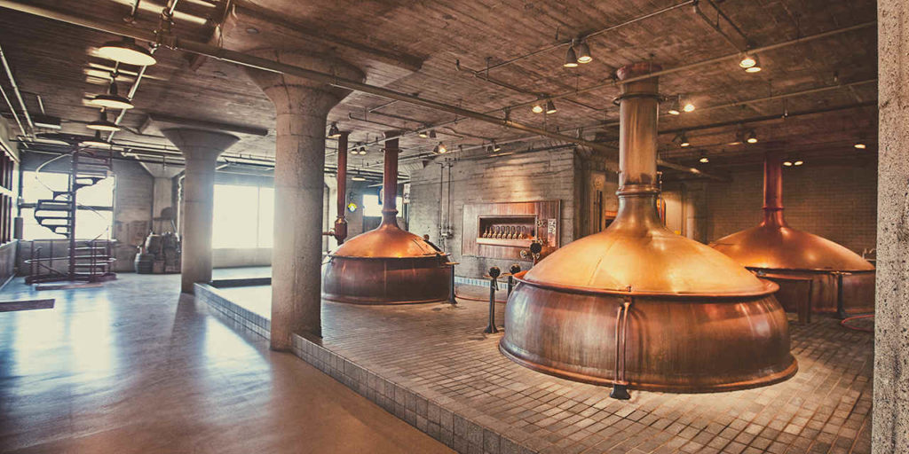 The image shows the interior of the Anchor Brewing Company's brewhouse, with several large metal brewing tanks in the foreground and background. The tanks are connected by pipes and other equipment, and there are stairs and walkways allowing access to the top of the tanks. The brewhouse has a rustic, industrial feel with exposed brick walls and metal beams. Several workers in green shirts and black pants are visible in the background, attending to the equipment. The Anchor Brewing Company logo is visible on a banner in the background.
